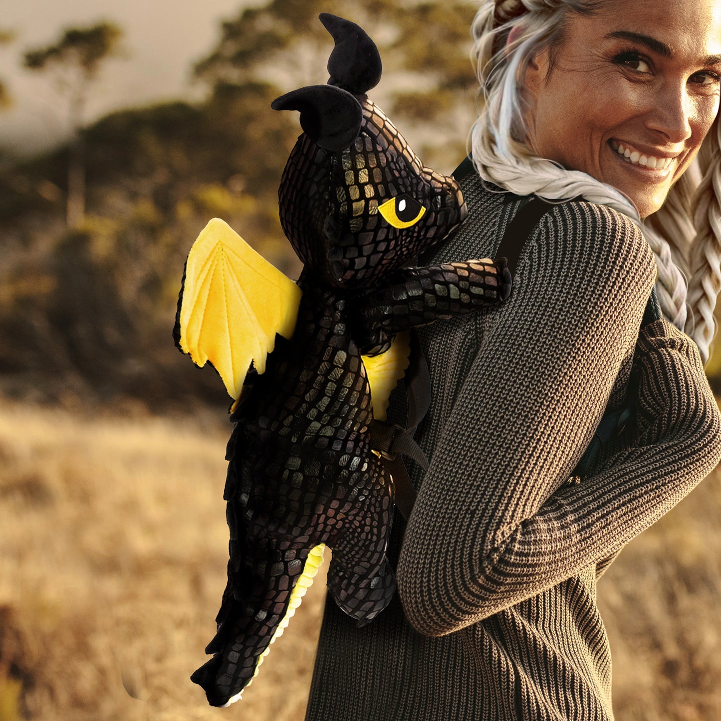 A female model in a grey sweater, standing in a field, wearing a black and yellow dragon-shaped backpack on her back.