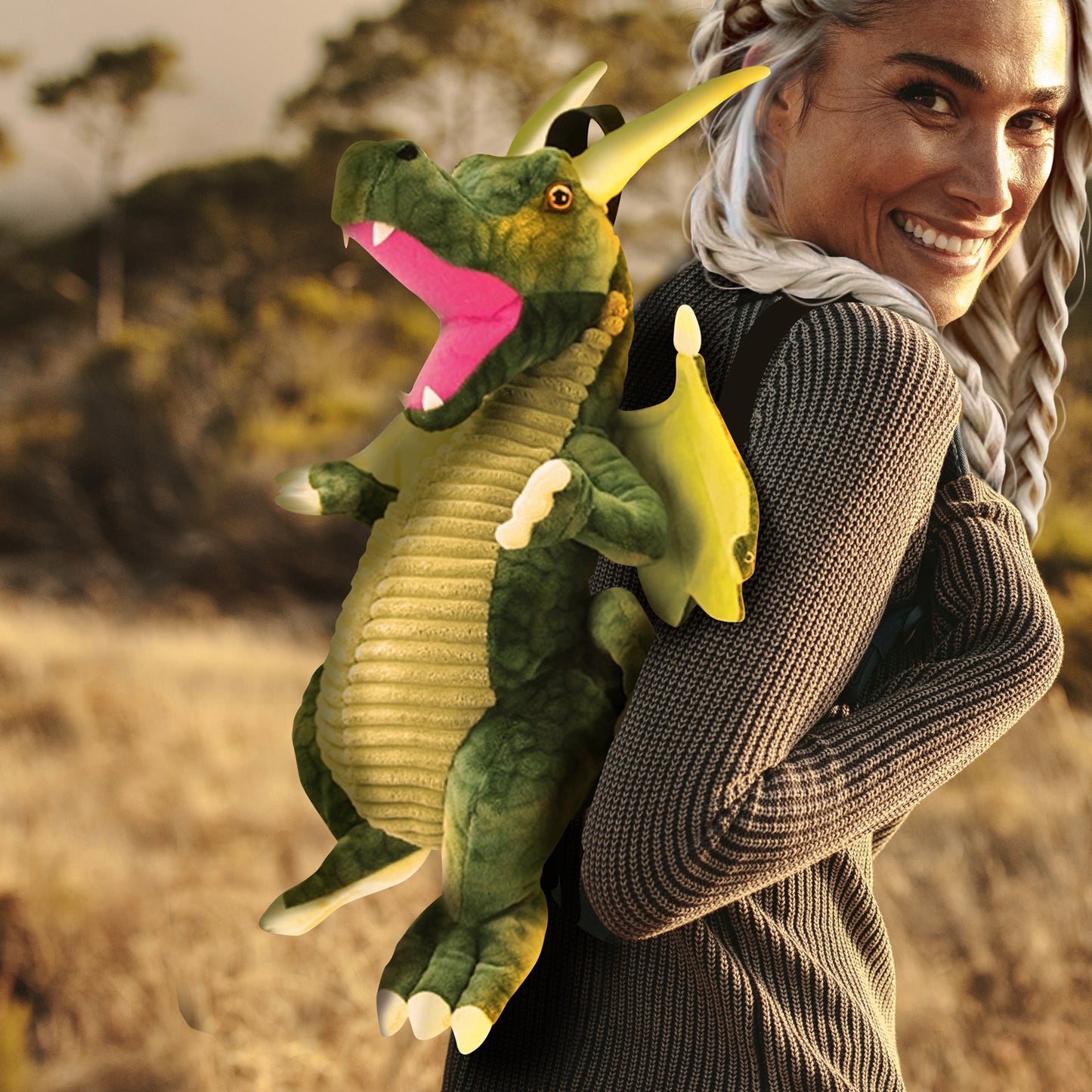 A female model wearing a green dragon-shaped backpack on her shoulders. The dragon has black carrying straps on its back. The model is in a field, with trees in the background.