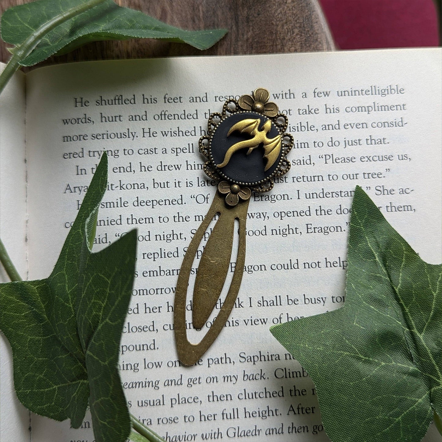 Decorative bookmark with a dragon design on an open book surrounded by green leaves.