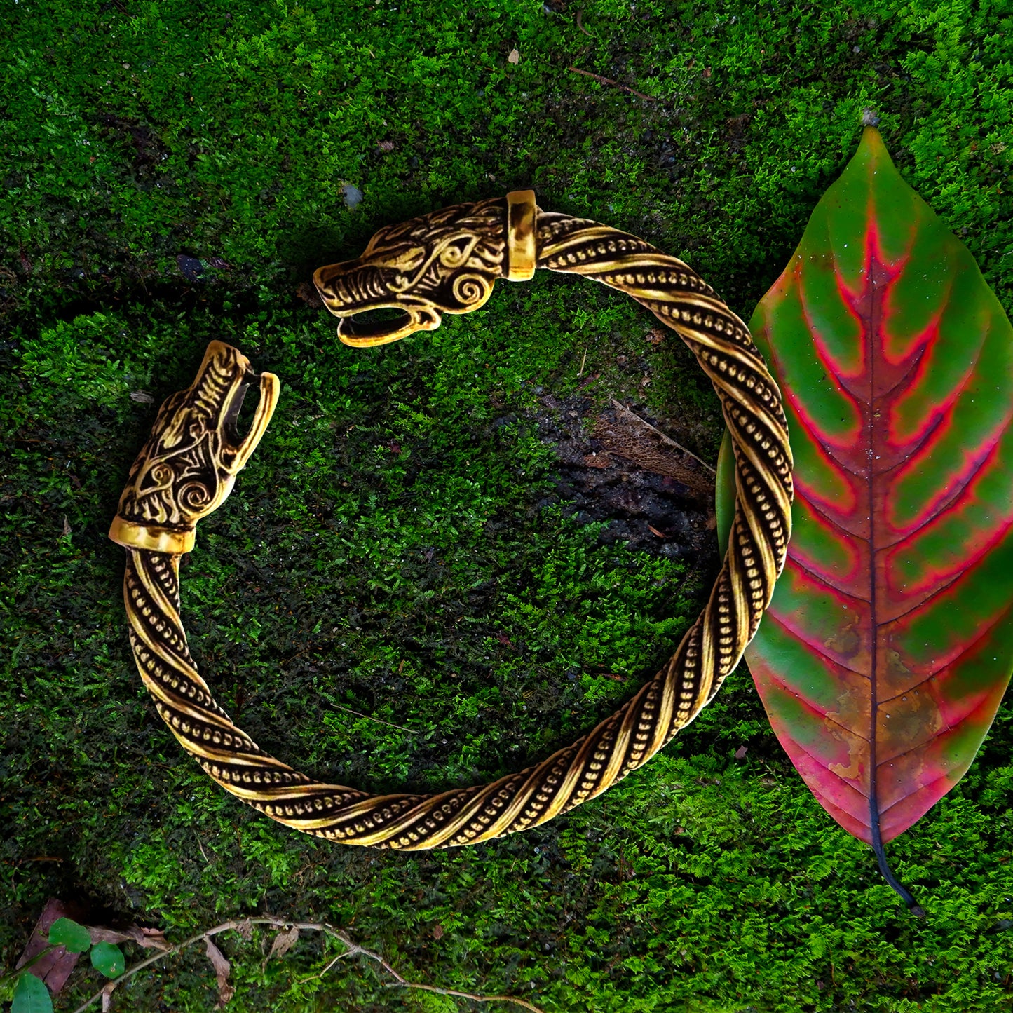 Close up view of a gold bracelet against a grassy background. Both ends of the bracelet have dragon heads on them, facing each other. The bracelet's main section has a spiraling line and dot pattern. Next to the bracelet is a green leaf with a red center.