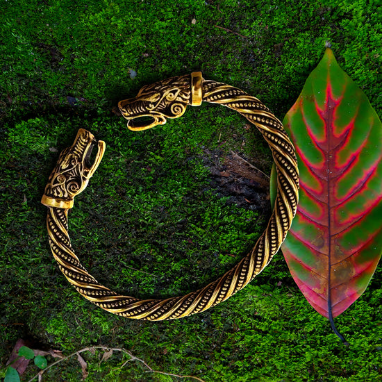 Close up view of a gold bracelet against a grassy background. Both ends of the bracelet have dragon heads on them, facing each other. The bracelet's main section has a spiraling line and dot pattern. Next to the bracelet is a green leaf with a red center.