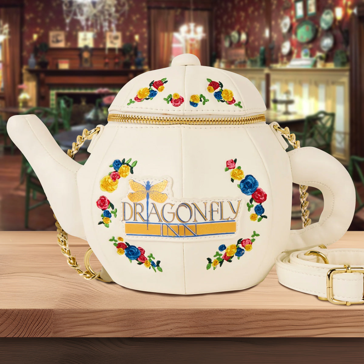 A white purse on a wooden table. The purse is shaped like a teapot. Around the front are colorful flowers. Gold and blue text says "Dragonfly Inn," with a gold and blue dragonfly next to it. The top of the teapot has a brass zipper. Next to the purse is a white strap, and a gold chain is attached to the front. In the background is an old fashioned dining room.