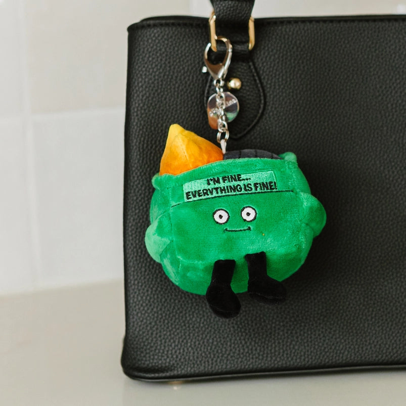 A green, square plushie depicting a dumpster on fire. The plushie has large white eyes and small black legs. Black text at the top says "I'm fine. Everything is fine!" A metal chain is attached at the top. The plushie is hanging from the strap of a black leather purse.