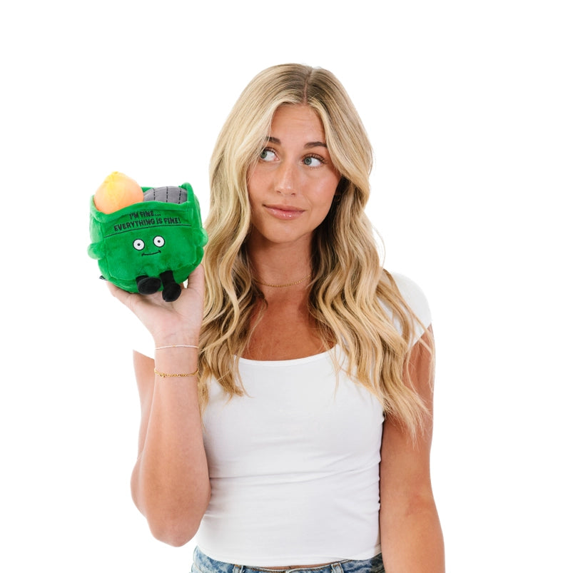 A female model holding a green, square plushie depicting a dumpster on fire. The plushie has large white eyes and small black legs. Black text at the top says "I'm fine. Everything is fine!"