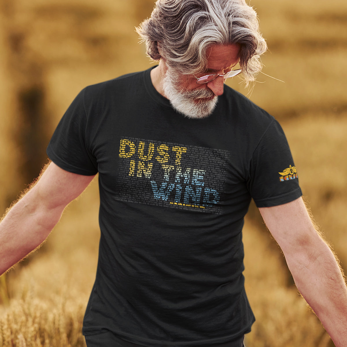 A male model wearing a black t-shirt standing in a wheat field. On the front of the shirt is text saying "dust in the wind." The text is comprised of smaller text, colored yellow and blue, to form the dust in the wind phrase. On the left sleeve is the logo of the band Kansas.