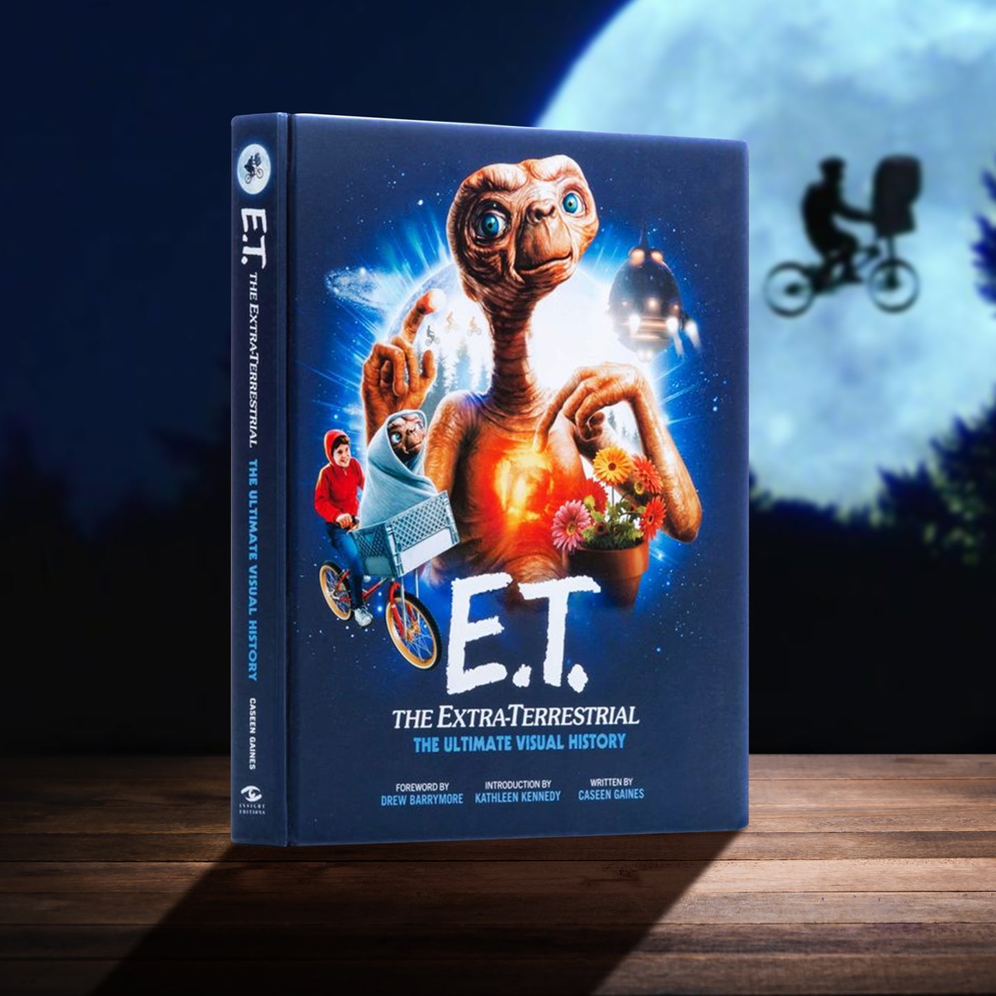 Book titled 'E.T. The Extra-Terrestrial: The Ultimate Visual History' with a cover featuring E.T. and a child on a bike. The book is on a wooden table. In the background is a full moon with a bicycle flying across the front.