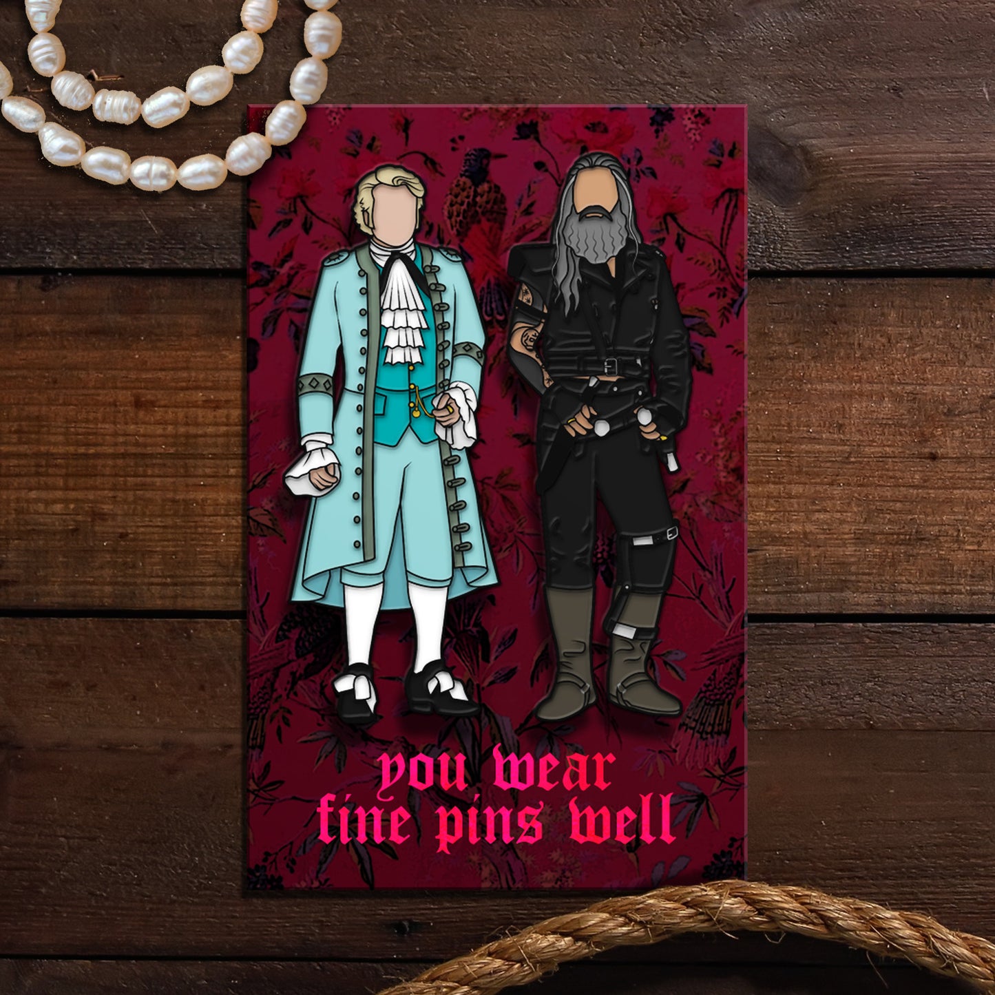 A pair of enamel pins on a red card. The left pin depicts a gentleman in light blue finery from the late 1600s. The right pin depicts a bearded man in dark pirate clothes. Red text at the bottom says "You wear fine things well." The background of the card has a pattern of birds and flowers. In the background is a wooden table with rope and pearls in the corners.