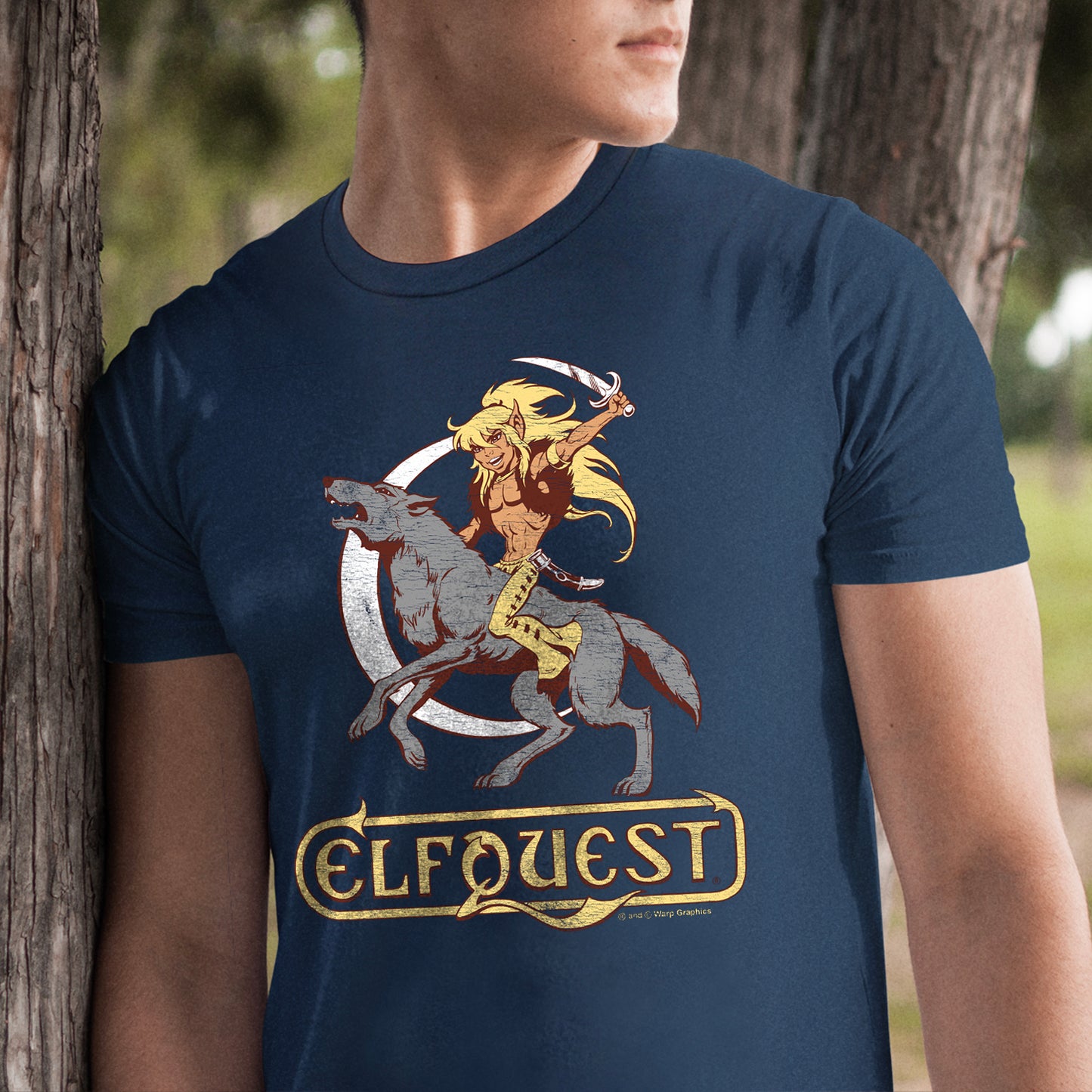 A male model leaning against a tree, wearing a blue t-shirt. On the front is a drawing of the character Cutter from the ElfQuest comic series, riding on a giant grey wold and brandishing a curved dagger. In the background is a narrow, white sliver of the moon. 