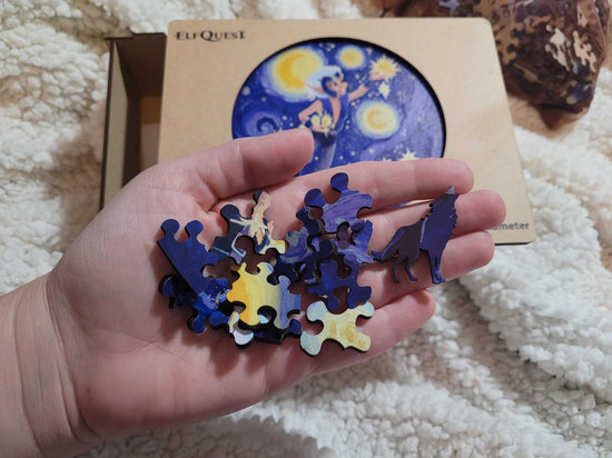 A model's hand holding blue and yellow puzzle pieces. In the background is a wooden box on a white fuzzy blanket. In the center is an image depicting the ElfQuest character Skywise in the sky, gathering stars.