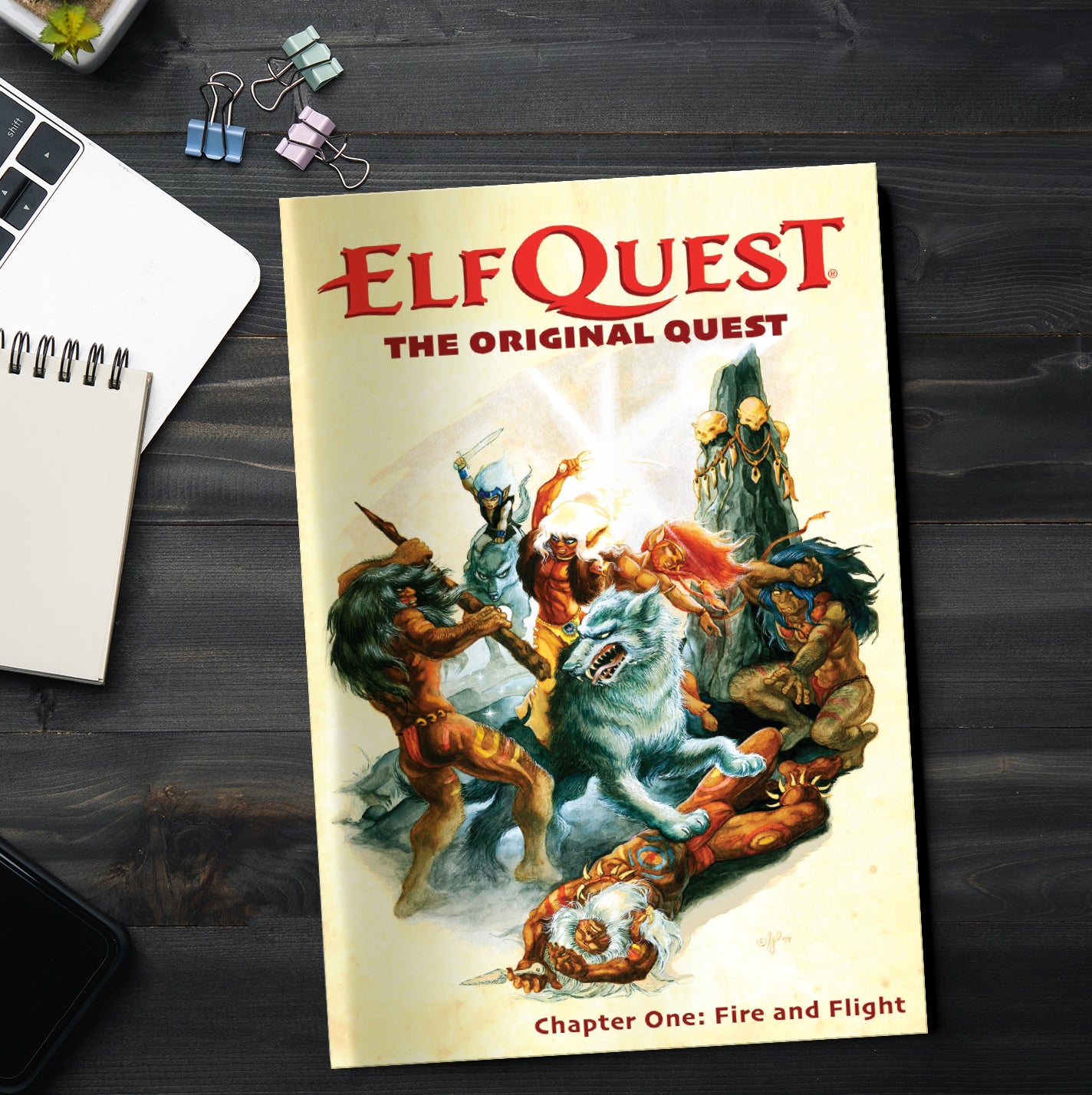 A copy of the comic book ElfQuest issue number one, on a wood desk. The cover depicts various armed elves in combat. Next to the comis is a laptop computer, a notepad, a cell phone, and paperclips.