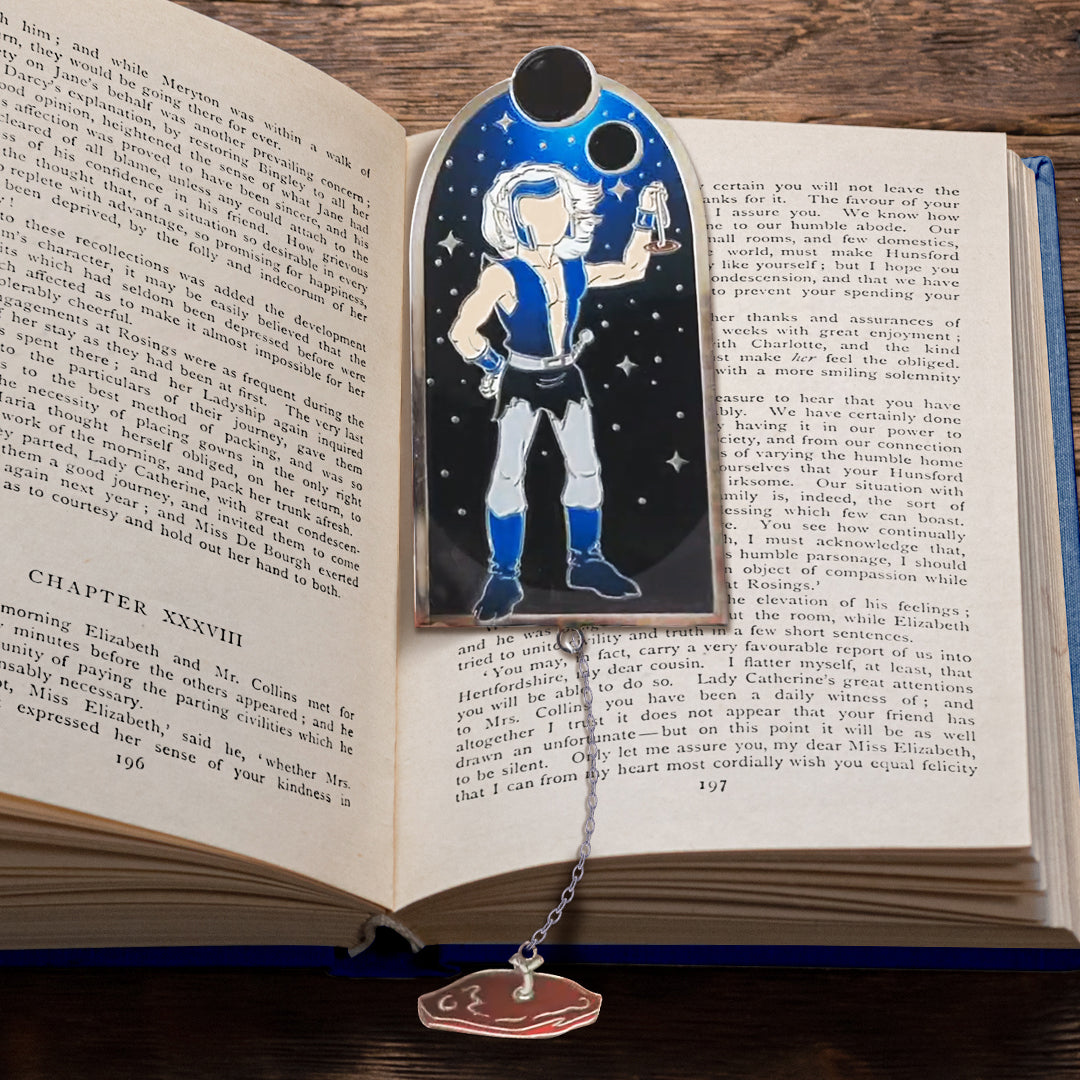 A metal bookmark sitting between pages of an open hardcover book, against a wooden table. The bookmark depicts an elf with long blonde hair, wearing a blue vest and black shorts. Behind him is a dark sky with stars scattered across, and two black moons at the top. A chain is attached to the bottom of the bookmark, with a brown charm at the end.