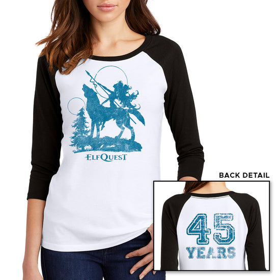 Two images of a female model wearing a white long sleeve T-shirt with black sleeves. The larger image has a blue image of an elf riding on the back of a large howling wolf, next to a small tree. The elf is holding a spear, and pointing it toward the sky. Under the wolf is text saying "ELFQUEST." The smaller image shows the back of the shirt, with "45 Years" printed in large blue text.