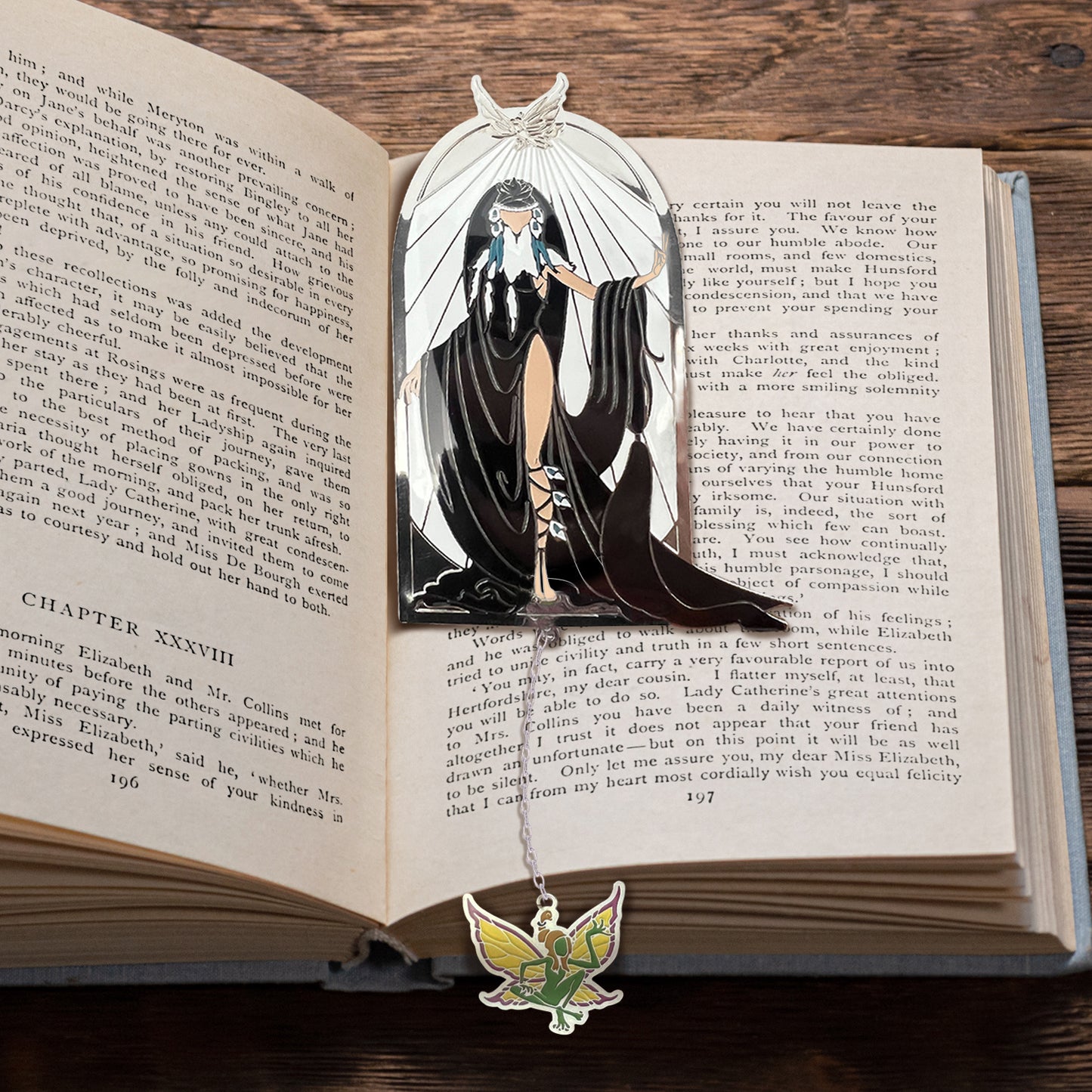 Close up view of a bookmark on the pages of a hardcover book. The bookmark depicts the ElfQuest character Winnowill, dressed in black robes with white rays above her. A chain hangs down from the bottom of the bookmark, and a green and yellow charm shaped like a fairy is attached to the end.