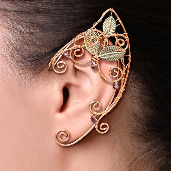 Close up of a model's ear, wearing a copper jewelry-wire ear cuff in the shape of an elven ear. It has swirling accents, dusty purple gems, and three green metal leaves, giving a woodland effect.