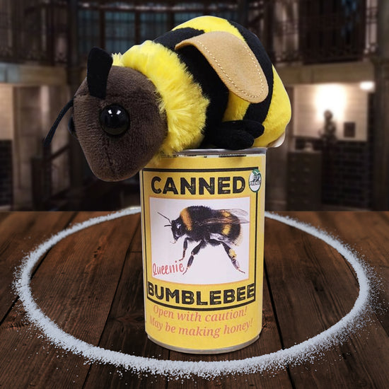 A bumblebee plushie sitting on a yellow can, on a wooden table. The back of the can has a white label, with bee-related jokes and facts in black and red text. A circle of salt surrounds the plushie and can. In the background is The Bunker.