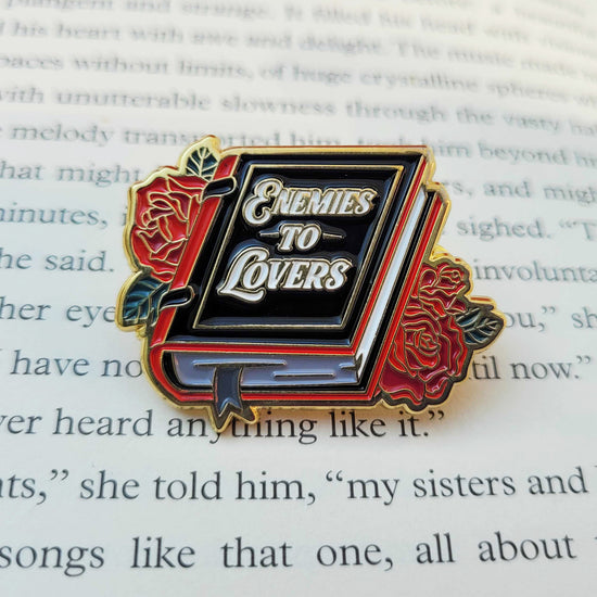 A black enamel pin on a book page. The pin is shaped like a book, with roses on each side, White text on the cover says "enemies to lovers."