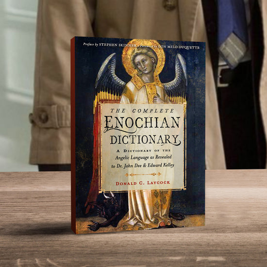 An image of the Complete Enochian Dictionary book cover, on a wooden tabletop with a tan trench coat hanging in the background. On the cover is a painting of an angel with a gold halo over her head. In front of the angel is a faint white square, with black text saying “The complete Enochian Dictionary: a dictionary of the angelic language as revealed to Dr. John Dee and Edward Kelley.” At the bottom of the book is the author’s name, Donald C Laycock, in red text.