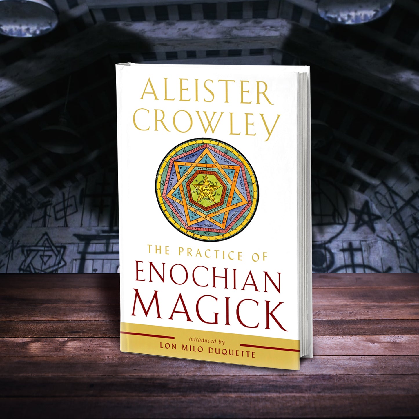 Load image into Gallery viewer, A white book on a wood table, set against a wall covered in magical symbols. At the top of the book in yellow text is the author&amp;#39;s name, Aleister Crowley. Under the name is a muilti-colored Enochian symbol, consisting of a six point star set within concentric circles. Under the symbol in yellow text is the title &amp;quot;The practice of Enochian Magick.&amp;quot; At the bottom of the cover is a yellow band, with red text reading &amp;quot;introduced by Lon Milo Duquette.&amp;quot;
