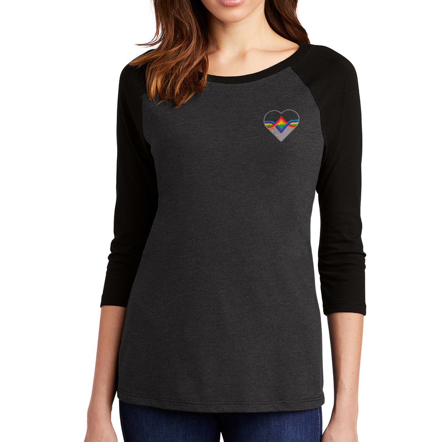 A female model wearing a grey longsleeve shirt with black sleeves. The Acting Ensign Pride symbol is at the top left corner of the shirt.  The symbol is a grey heart with rainbow lines running through the center.