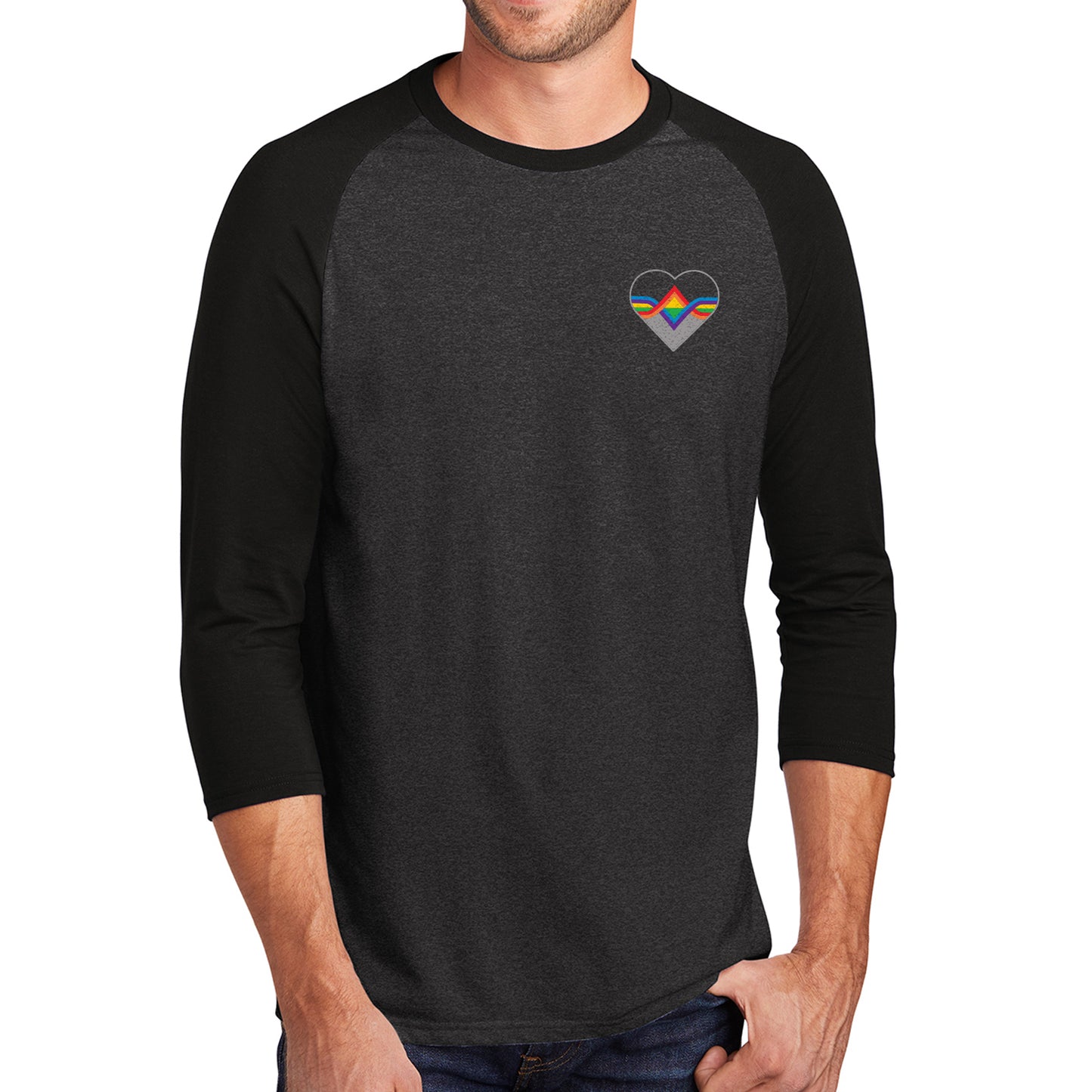 A male model wearing a grey longsleeve shirt with black sleeves. The Acting Ensign Pride symbol is at the top left corner of the shirt.  The symbol is a grey heart with rainbow lines running through the center.