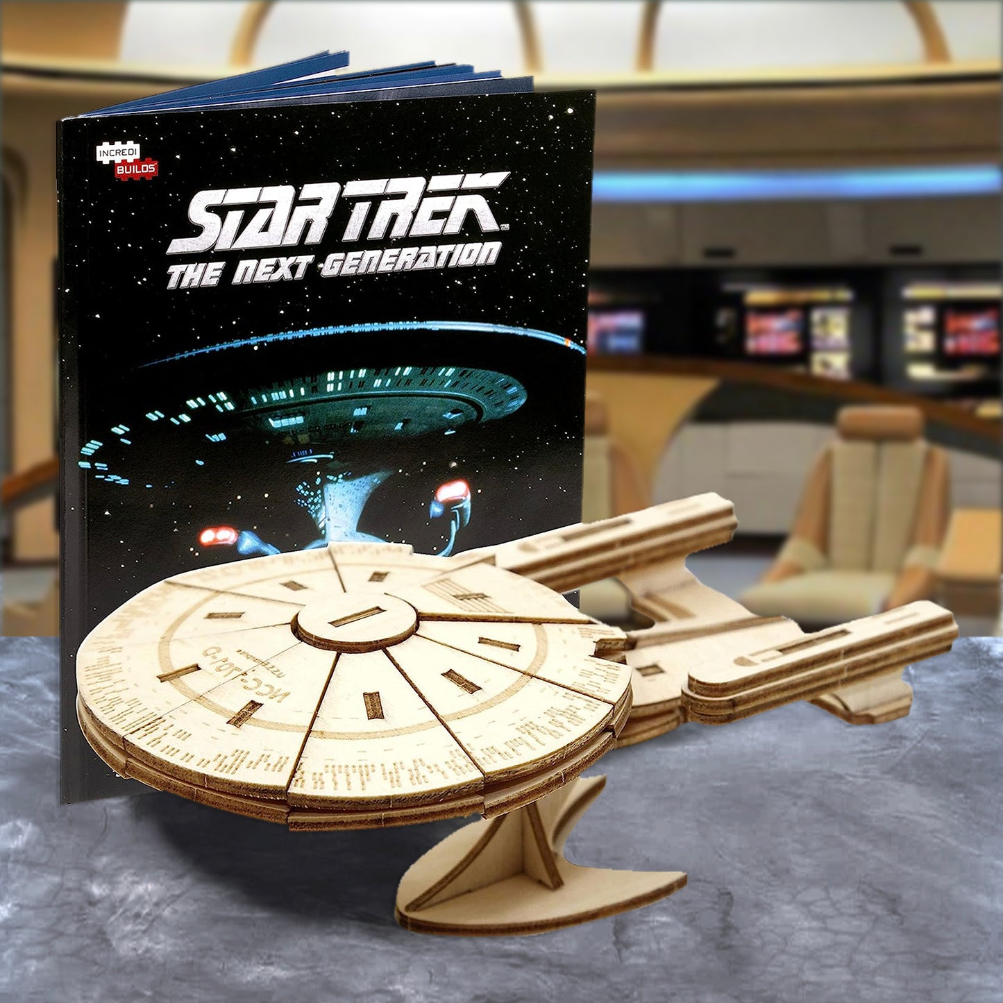 A wooden model of the Starship Enterprise, NCC-1701-D, from Star Trek The Next Generation. Next to the model is a guide book for assembly instructions. In the background is the bridge of the Enterprise.