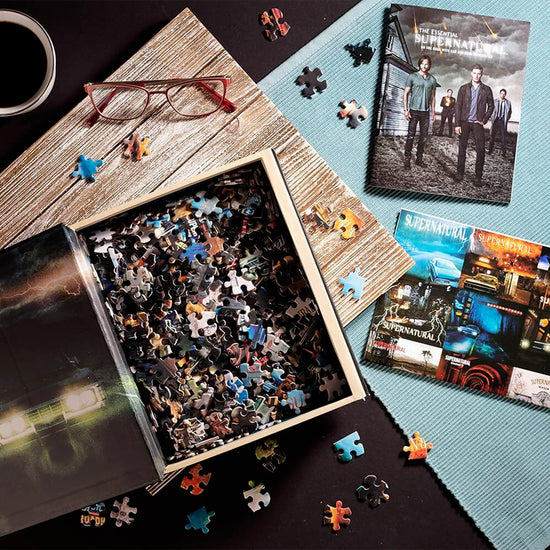 An image of a book on a tabletop, opened to reveal a hollow space filled with jigsaw puzzle pieces. On the inside cover is an image of a 1967 Impala on a dark road, underneath lightning bolts in the sky. Surrounding the book are a blue placemat, a cup of coffee, reading glasses, and a softcover book with an image of characters from Supernatural on the cover.