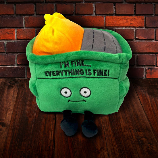 A green, square plushie depicting a dumpster on fire, sitting on a dark wooden floor. The plushie has large white eyes and small black legs. Black text at the top says "I'm fine. Everything is fine!" In the background is a brick wall.