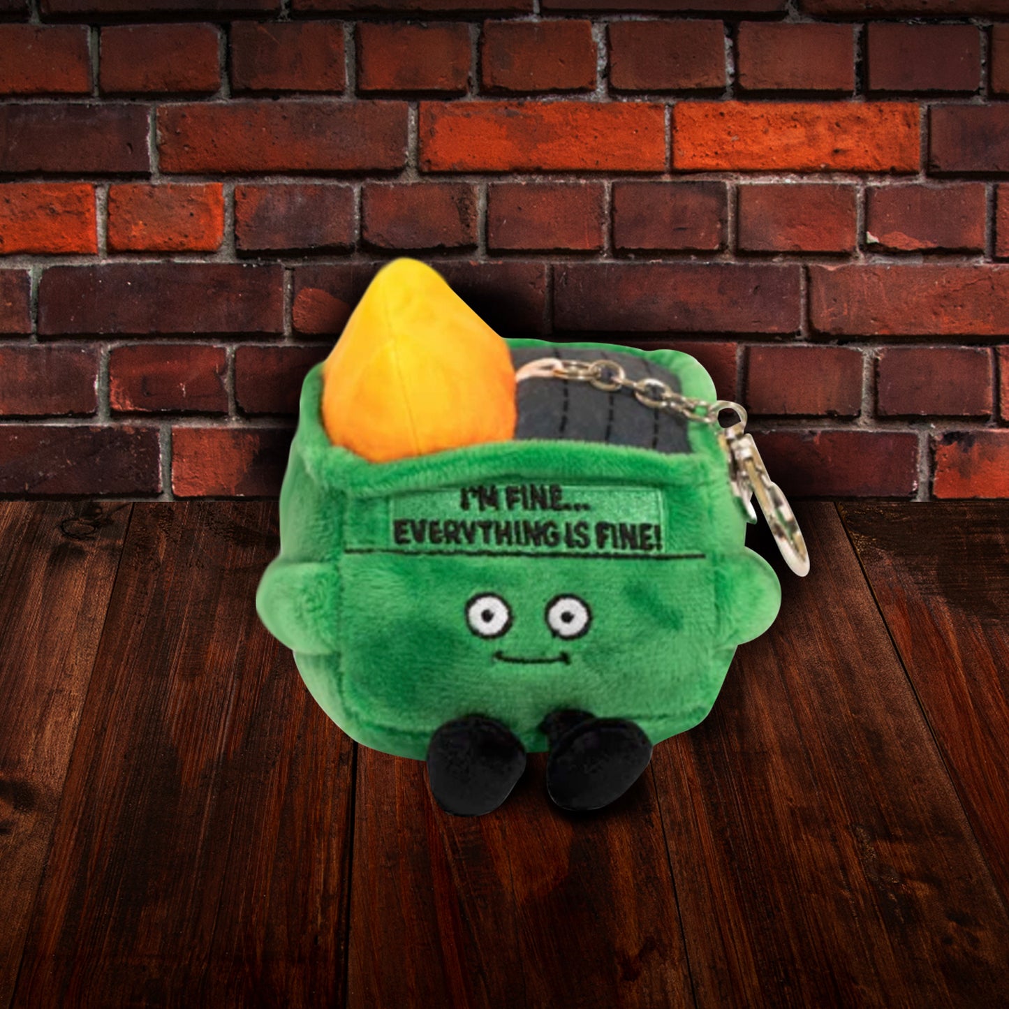 A green, square plushie depicting a dumpster on fire, sitting on a dark wooden floor. The plushie has large white eyes and small black legs. Black text at the top says "I'm fine. Everything is fine!" A metal chain is attached at the top. In the background is a brick wall.