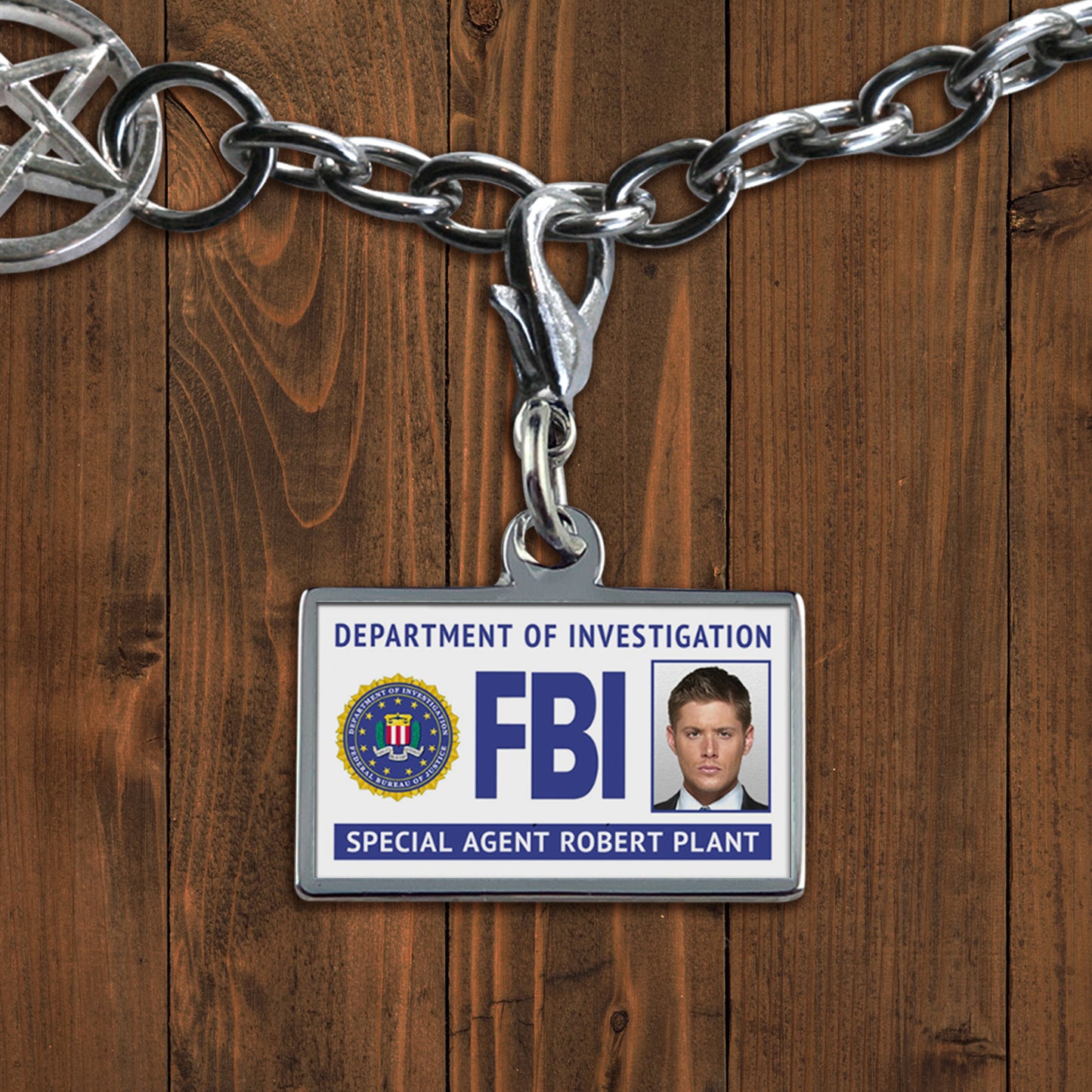 A white charm against a wooden background. The charm depicts an FBI badge, with Castiel's face at the side. At the bottom is white text saying "Special agent Eddie Moscone." The badge is upside down.