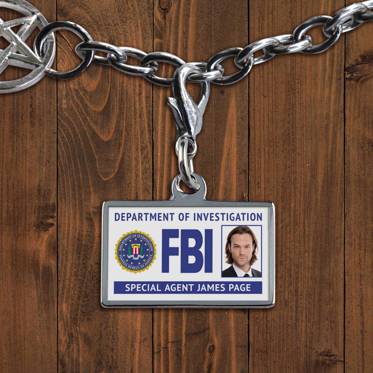 A white charm against a wooden background. The charm depicts an FBI badge, with Dean Winchester's face at the side. At the bottom is white text saying "Special agent Robert Plant."