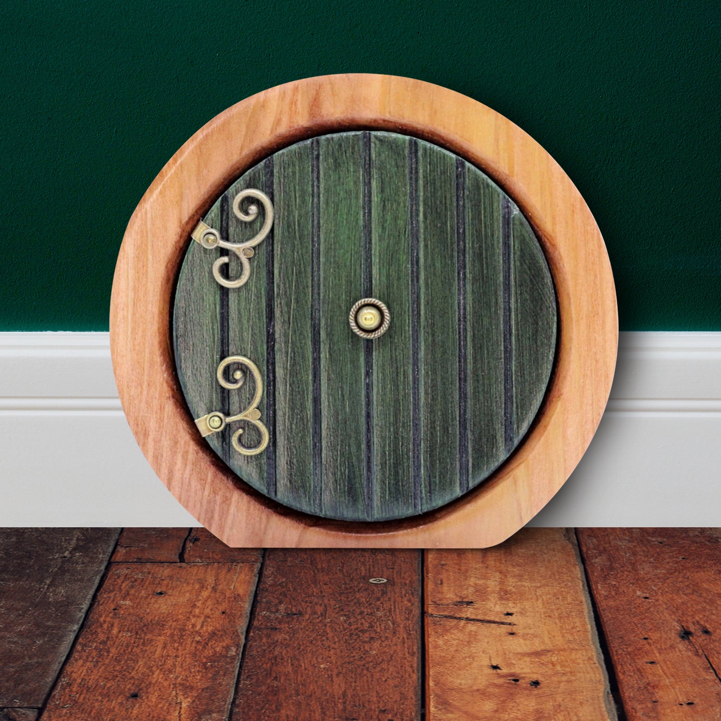 A round wooden doorframe with a green door on a wooden floor, against a white baseboard and green wall. The door has ornate brass hinges at the side, and a brass doorknob in the center. 