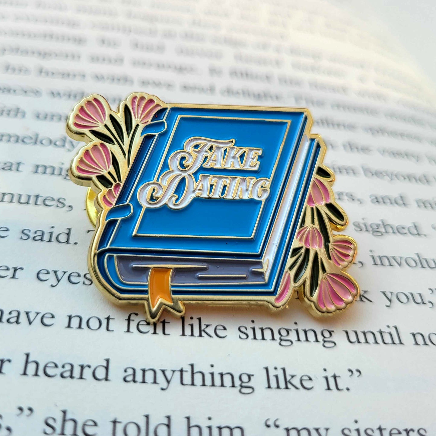 A blue enamel pin on a book page. The pin is shaped like a book, with pink flowers on on each side, White text on the cover says "fake dating."