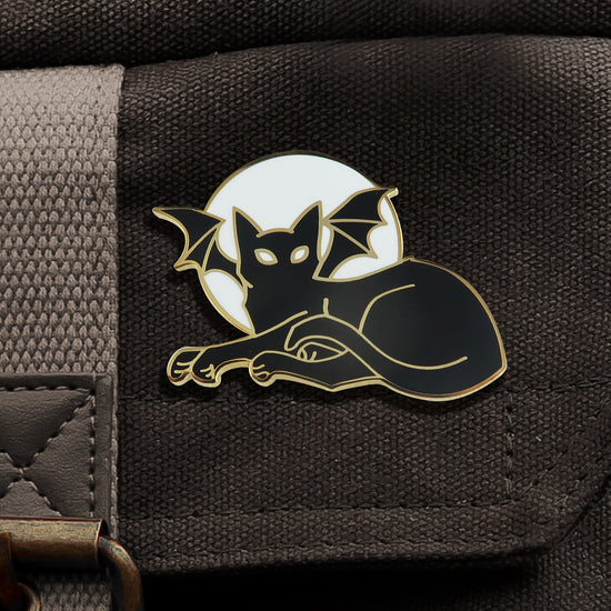 A black enamel pin with gold edges, attached to a black canvas bag. The pin depicts a lounging black cat with bat wings. Behind the cat's head is a round white moon.