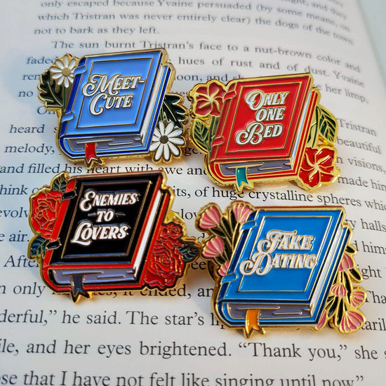 Four book-shaped enamel pins against a book page. Each pin is a different color, with gold text on the cover spelling out a romantic trope. Only One Bed, Meet Cute, Fake Dating, Enemies To Lovers.