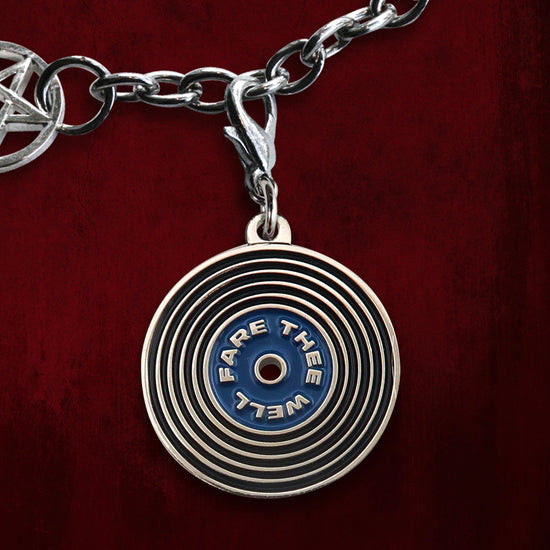 A silver charm hanging from a silver charm bracelet and set against a dark red background. The charm resembles a record with a blue inner ring, which reads "Fare Thee Well" within the ring.