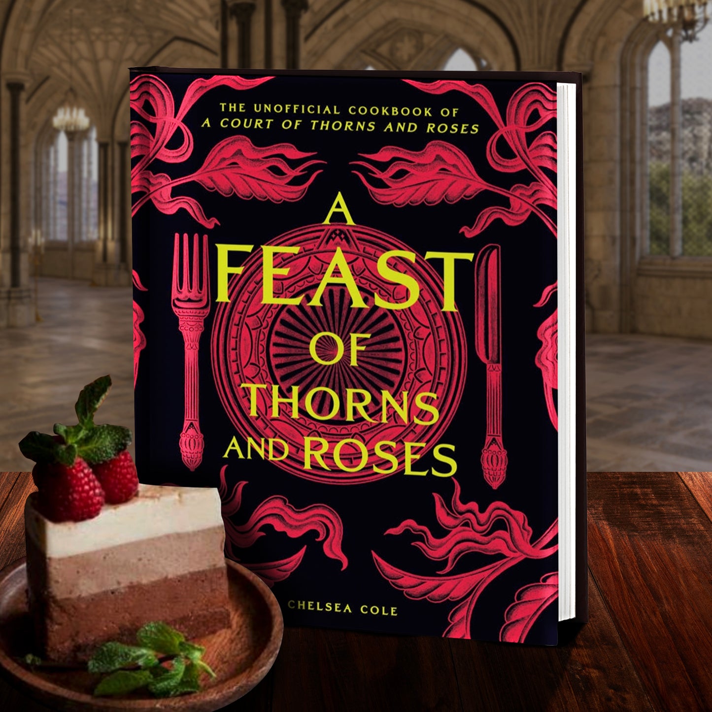 A red and black hardcover book on on a wooden table. The cover depicts a red and black image of a fancy dinner table setting. Yellow text says "The unofficial cookbook of a court of thorns and roses. A Feast of thorns and roses." Next to the book is a three layered dessert on a wooden plate. In the background is a castle courtyard.