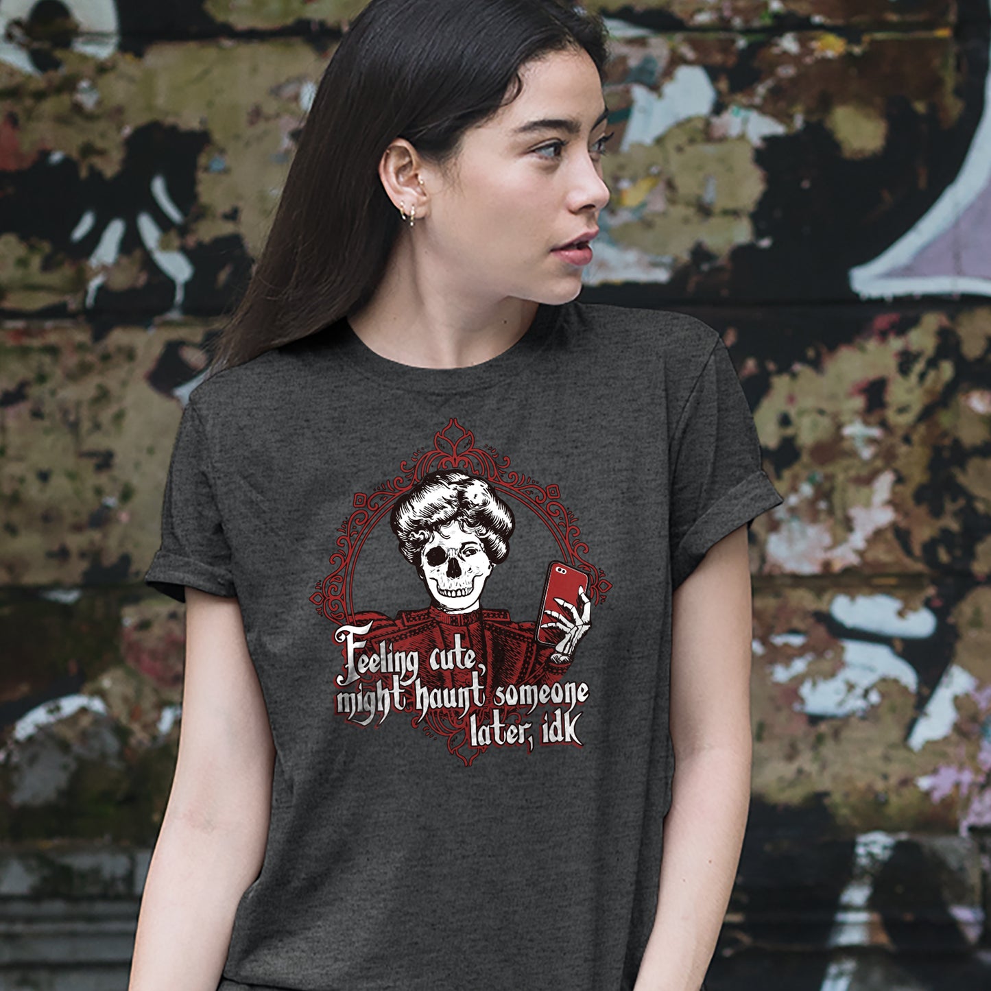 A female model wearing a grey T-shirt, standing against a wall with peeling paint. On the shirt is a skeletal figure holding a red cell phone, surrounded by a red filigreed border. Under the figure in white text is "feeling cute, might haunt someone later, IDK."