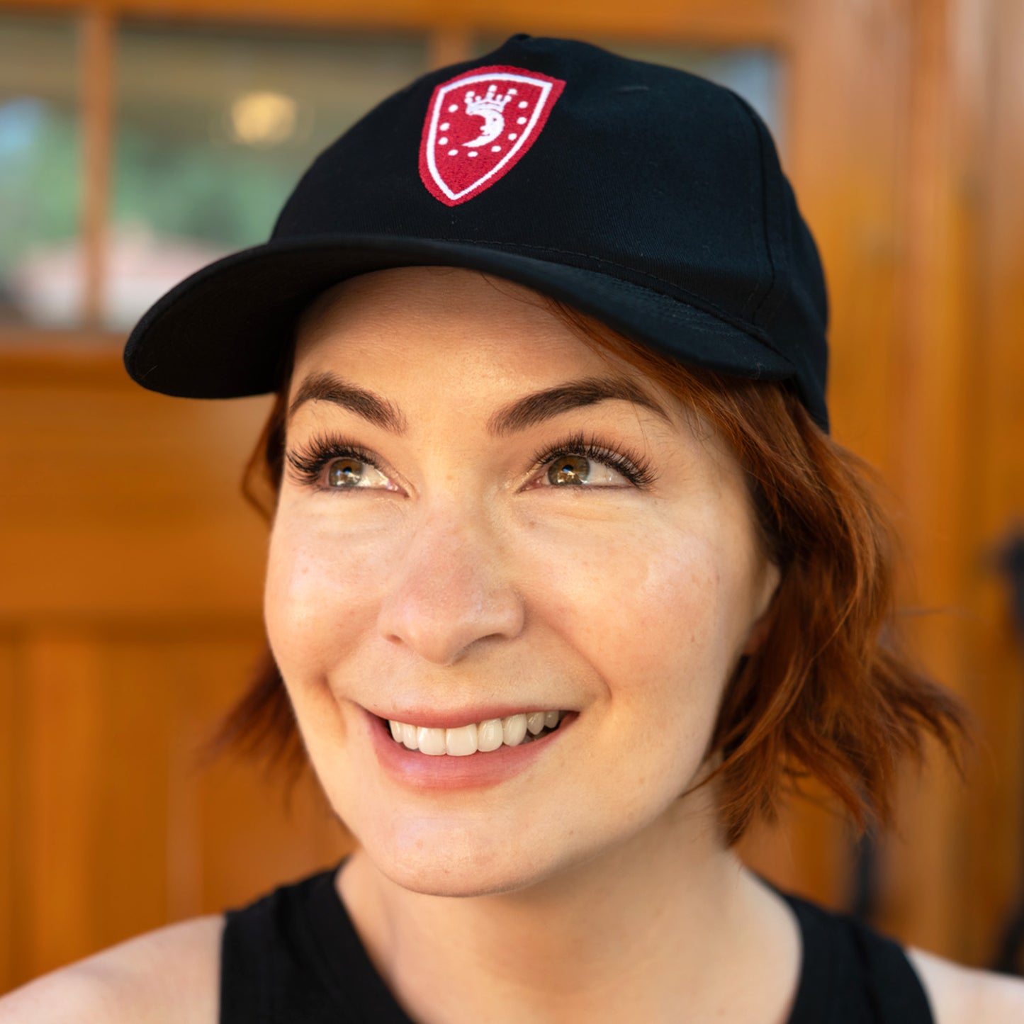An image of actor Felicia Day wearing a black cap with a red and white emblem. The emblem depicts a white crescent moon wearing a crown, with white dots around it in a circle, against a red shield-shaped background.