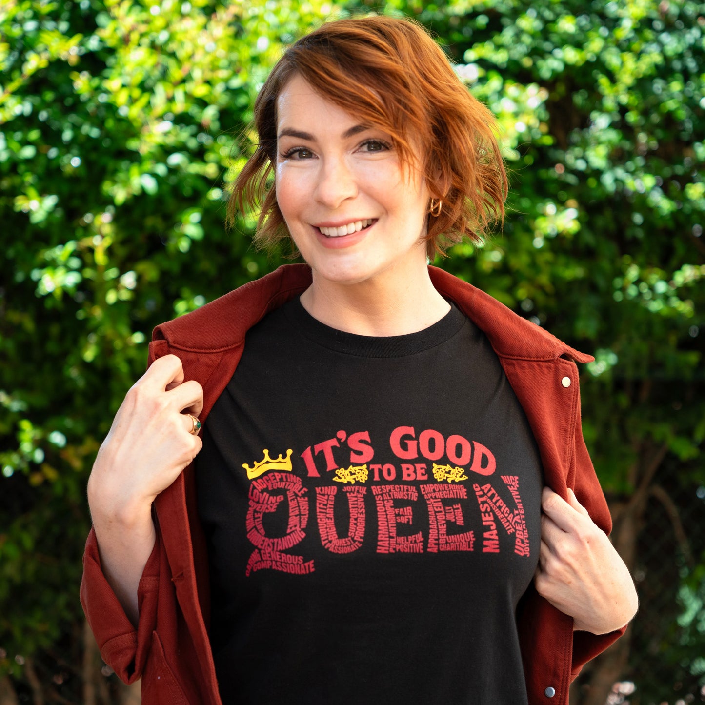 An image of actor Felicia Day wearing a black tee shirt. On the front of the shirt is red text saying "it's good to be queen," with a yellow crown on the Q. The letters in "queen" are made up of royal-themed words.