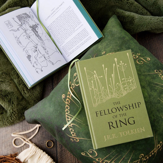 Two green hardcover books on a green pillow. The front cover of one book depicts a minimalist drawing of a swamp in gold. Black text says "The Fellowship of the Rings." The second book is open to show a map depicting an area in Middle Earth.