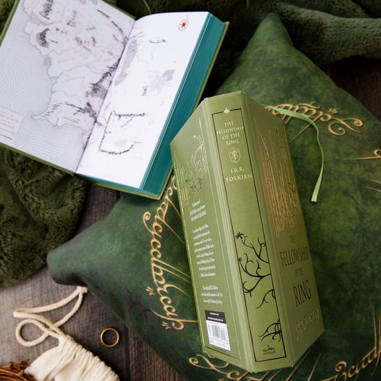 Two green hardcover books on a green pillow. The front cover of one book depicts a minimalist drawing of a swamp in gold. Black text says "The Fellowship of the Rings." The second book is open to show a map depicting an area in Middle Earth.