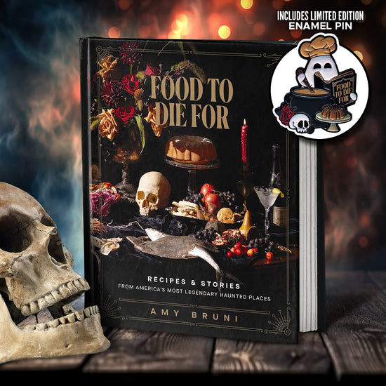 A black book on a wooden table. On the front cover of the book are berries, candles, cakes, and flowers, against a black background. Gold text at the top says "Food to die for." At the bottom is white text saying "recipes & stories from america's most legendary haunted places." Next to the book is a human skull. In the background is smoke and sparks. At the top right is an enamel pin, shaped like a ghost holding the book in one hand while stirring a cauldron with the other.