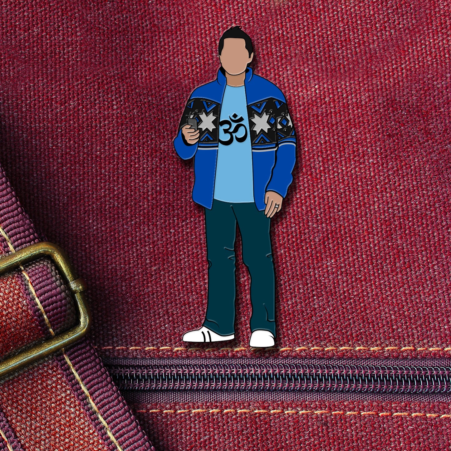 An enamel pin depicting Castiel from the TV series "Supernatural." Castiel is a blue shirt with a yoga symbol on it, and a dark blue sweater over it. In one hand is his cell phone. The pin is attached to a maroon canvas bag.
