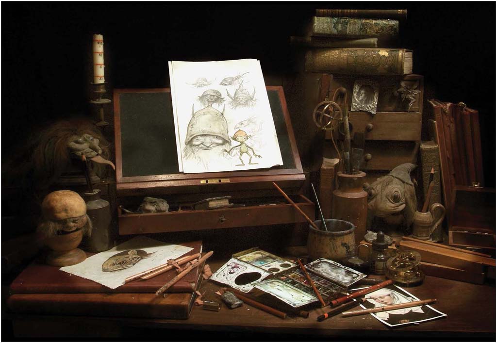 A two-page spread from the book depicting an old-fashioned illustrator's desk, with various colored pencils and paints. Wooden drawers and models are strewn across the workspace, and at the center is a white page with sketches of faerie drawings.