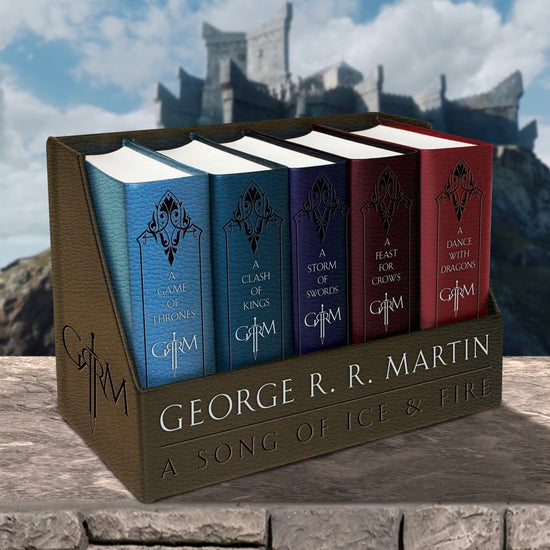 Close up of a boxed set of George R. R. Martin's "A Song Of Ice & Fire" book series, sitting on a stone wall. Behind the book set is a castle on a mountain top.