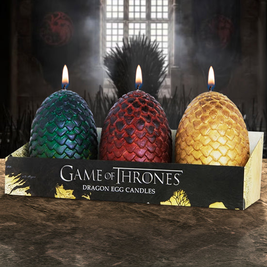 Three egg-shaped candles in a cardboard package, on a stone table. The eggs have scales around their outside. One is green, one is red, and one is gold. The package is black and gold, with silver text that says "Game of Thrones, dragon egg candles." The candles are all lit. In the background is a castle room with stone walls and a stained glass window