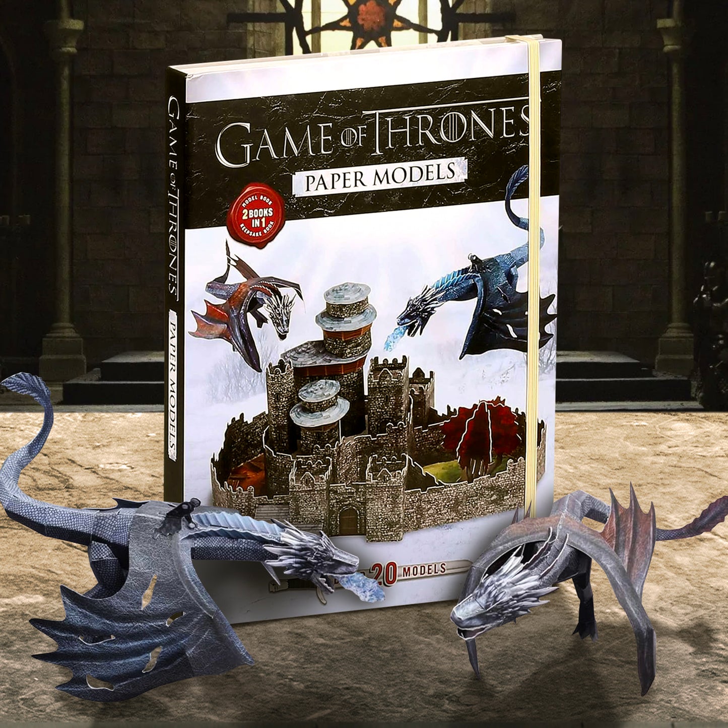 A cardboard box on a stone table. The cover depicts a scene from the TV series Game of Thrones, with two paper model dragons attacking a paper model castle. In front of the box are two paper model dragons, one black and one red. White text says "Game of Thrones paper models."