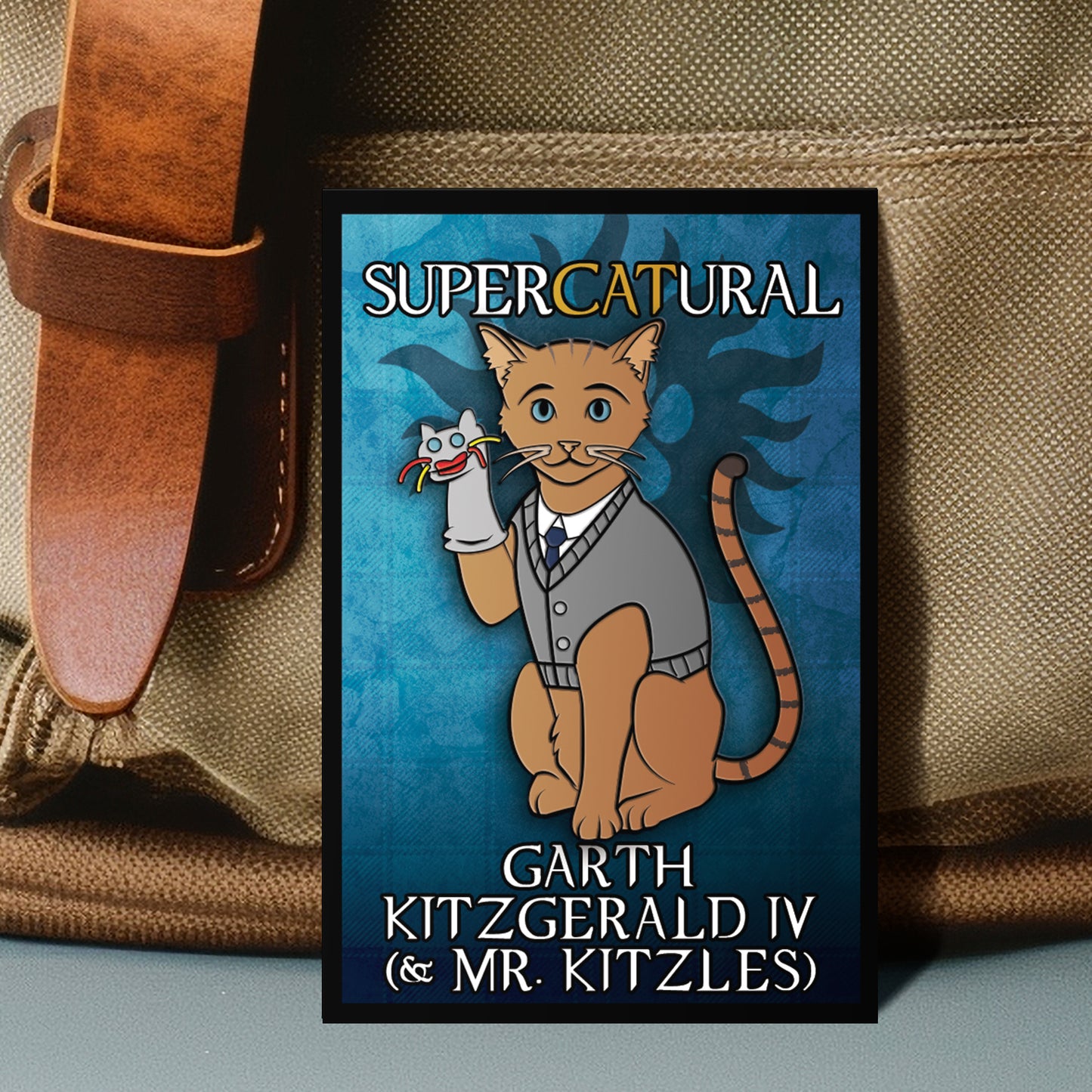 Poster of a cat character with text 'Supercatural' and 'Garth Kitzgeral IV & Mr. Kitzles' on a blue background, leaning against a brown bag.