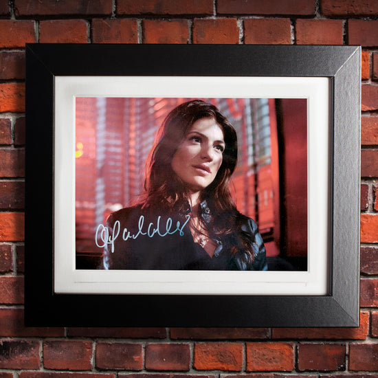 A black framed photograph against a brick wall. The photograph depicts actor Genevieve Padalecki, dressed as the character Ruby from the series "Supernatural." Gen is standing in front of windows with red-lit blinds, wearing a black leather jacket. In the bottom left corner is Gen's autograph in white ink.