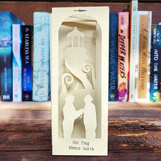 A white paper bookmark on a wooden table set against a bookshelf background. The bookmark depicts a lighthouse, and silhouette cut-outs of characters from the series "Our flag means death." In front of the lighthouse are kraken tentacles. Silver text at the bottom of the bookmark says "our flag means death."